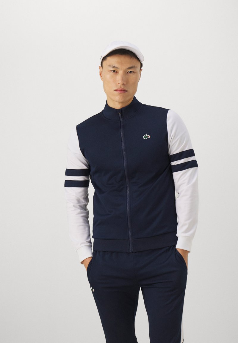 Veste de survêtement zippée marine avec des manches longues blanches et des accents rayés. Présente un logo vert sur la poitrine et un design de col classique.