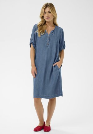 Džinsinė suknelė su apvalia iškirpte, sagų sąsašu, trumpomis susuktais rankovėmis, šoninėmis kišenėmis ir laisvu fasonu. Dėvima su raudonais austais slip-on bateliais.