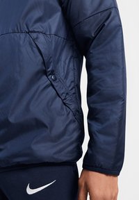 Marineblauwe gewatteerde jas met een gladde, glanzende textuur; heeft een met rits afsluitbare zijzak en elastische boorden voor een snugge pasvorm.