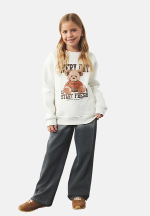 Sweatshirt blanc arborant un graphisme de ours en peluche marron, associé à un pantalon ample gris foncé et des baskets marron avec des lacets beiges.