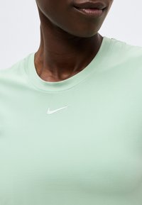 Κοντινό πλάνο ενός ατόμου που φοράει ανοιχτοπράσινη μπλούζα με στρογγυλή λαιμόκοψη και μικρό λευκό λογότυπο Nike τοποθετημένο στο κέντρο κάτω από τη λαιμόκοψη.