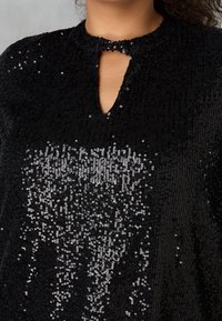 Top nero realizzato in tessuto lucido di paillettes, con un colletto alto e un piccolo dettaglio a chiave sul petto. Superficie testurizzata con accenti riflettenti.