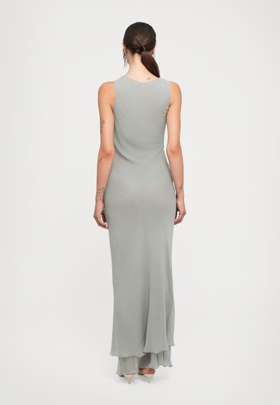 Woman with a low ponytail wearing a sleeveless, floor-length light gray dress and beige heels, standing with her back facing the viewer.