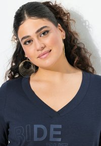 Camiseta de cuello en V de color navy con tejido texturizado; presenta la palabra "RIDE" en gris. El modelo lleva pendientes grandes de oro en diseño en capas.