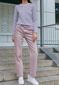 Randig långärmad skjorta i vit och lila med en liten orange logotyp; rosa byxor med sidofickor; vita sneakers med rosa accenter.