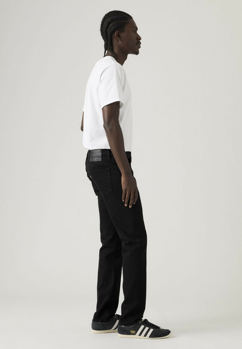 Mann mit geflochtenem Haar steht im Profil und trägt ein weißes T-Shirt, schwarze Jeans und schwarze Adidas-Sneaker vor einem schlichten hellen Hintergrund.