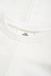 Balta kokvilnas T-krekls ar apaļu kakla izgriezumu un minimālu šuvju detaļām, ar redzamu etiķeti apkakles iekšpusē. Gluda virsma.