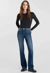 Donkerblauwe, uitlopende jeans met een medium wassing, ontwerp met vijf zakken en een knoopsluiting. Draagt een aansluitend zwart shirt met lange mouwen.