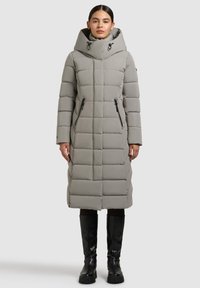 Lichtgrijze lange gewatteerde jas met capuchon, voorzien van een doorgestikt design, twee zijritsen en zwarte accenten, gedragen met kniehoge zwarte laarzen.