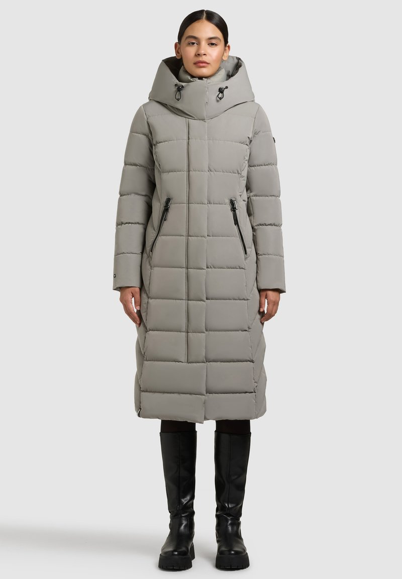 Lichtgrijze lange gewatteerde jas met capuchon, voorzien van een doorgestikt design, twee zijritsen en zwarte accenten, gedragen met kniehoge zwarte laarzen.