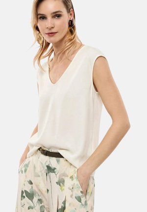 Donna che indossa una canottiera bianca con scollo a V, pantaloni a stampa floreale e una cintura scura, in piedi con le mani nelle tasche, sfondo chiaro.