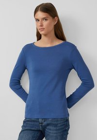 Junge Frau mit langen braunen Haaren, die ein einfaches blaues Langarmshirt und blaue Jeans trägt, steht vor einem weißen Hintergrund.