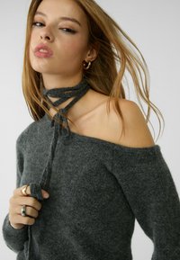 Grau, schulterfreier Pullover mit weicher Textur und gebundenem Ausschnitt. Die Accessoires bestehen aus mehreren goldenen Ringen und einem strukturierten Armband.
