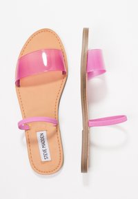 Des sandales plates roses avec une sangle transparente et un bracelet de cheville fin rose, présentant une semelle intérieure beige et une semelle de couleur marron clair.