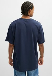 Hombre con trenzas en cornrow, vistiendo una camiseta holgada de color azul marino y pantalones cortos de mezclilla azul claro, de espaldas frente a un fondo liso.