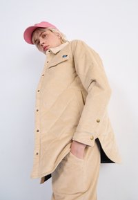 Beige kordfløyelsjakke med diamantmønster, lukket med gullsnapper, paret med matchende bukser og en rosa caps.