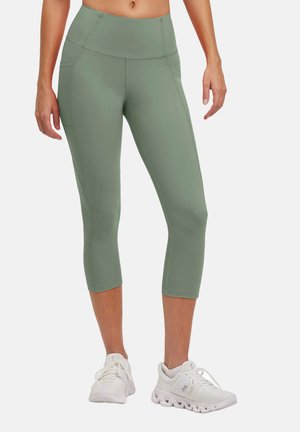 Green capri leggings made of stretch fabric, featuring a high waistband, side pockets, and seamless construction. Worn with white athletic shoes.