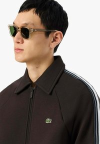 Chaqueta marrón con cremallera y cuello ancho, con un pequeño logo verde de Lacoste y rayas blancas en los laterales. El modelo lleva gafas de sol transparentes.