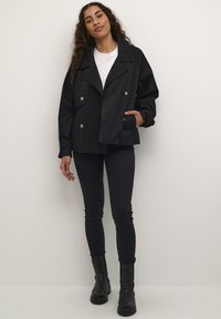 Svart dubbelknäppt jacka med stora knappar, breda slag och avslappnad passform. Bärs över en vit t-shirt och kombineras med svarta byxor och stövlar.