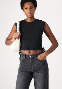Black cropped top with sleeveless design and side ties. Paired with high-waisted dark jeans and a cream shoulder bag. Smooth fabric texture.