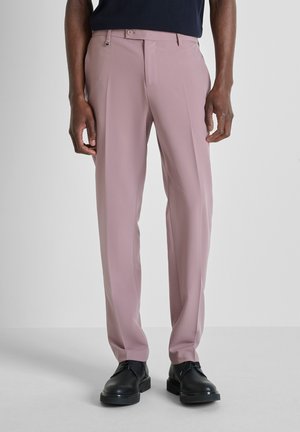 Uomo con pantaloni su misura di un tenue colore malva e scarpe eleganti in pelle nera, su uno sfondo chiaro e uniforme.