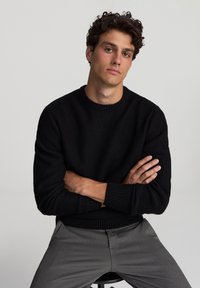 Maglione nero lavorato a maglia con scollo rotondo e polsini a costine. Il modello ha le braccia incrociate e indossa pantaloni grigi con texture. Sfondo neutro.