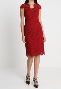 Robe en dentelle rouge jusqu'au genou avec des bords festonnés, des manches courtes et une silhouette ajustée, associée à des escarpins noirs à talons aiguilles.