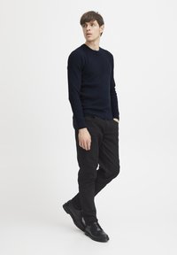 Pulover din tricot cu model cord, pantaloni negri și pantofi negri cu șireturi; modelul stă de profil cu mâinile în buzunare, fundal minimalist.