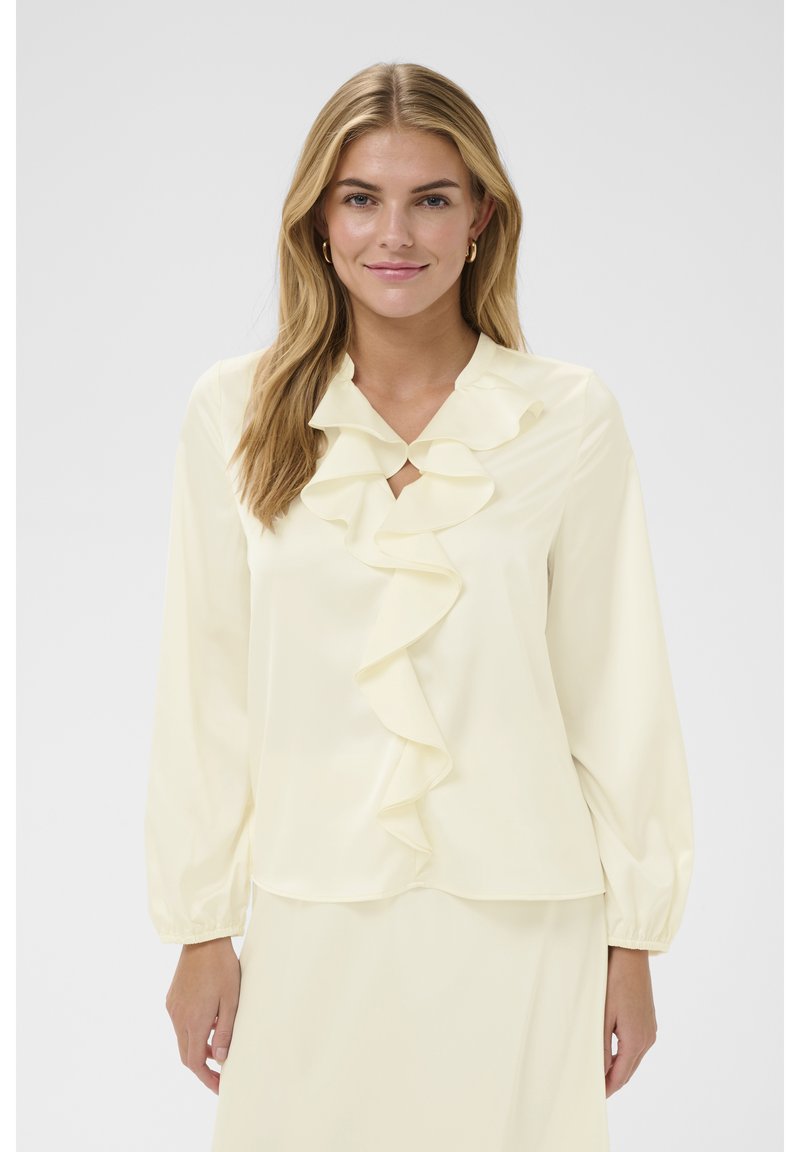 Jeune femme aux longs cheveux blonds portant un chemisier crème à volant sur le devant et manches longues, debout devant un fond clair uni.