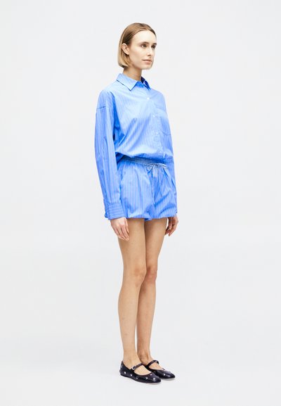 Γυναίκα στέκεται στραμμένη προς τα δεξιά φορώντας φαρδιά μπλε ριγέ πουκάμισα με μακριά μανίκια, ταιριαστά σορτς και μαύρα παπούτσια με λεπτές λουρίδες στον αστράγαλο.