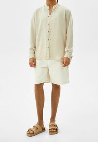 Beige overhemd met lange mouwen, witte korte broek en lichtbruine sandalen. Eenvoudig ontwerp met een ontspannen pasvorm en lichte stoftextuur.