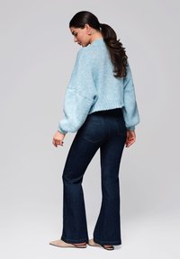 Ljusblå stickad kort sweater med breda ärmar, kombinerad med mörka utsvängda jeans. Modellen visar en sidovy mot en neutral bakgrund.