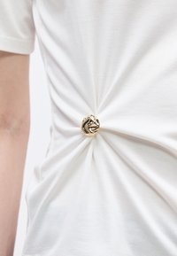 Λευκό πουκάμισο μαζεμένο και στερεωμένο στη μέση με ένα λαμπερό, ακανόνιστο χρυσό κουμπί, δημιουργώντας πτυχές υφάσματος που ακτινοβολούν προς τα έξω.