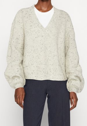 Personne portant un pull en maille crème tacheté avec un col en V et des manches longues bouffantes sur un haut blanc avec un pantalon gris foncé.