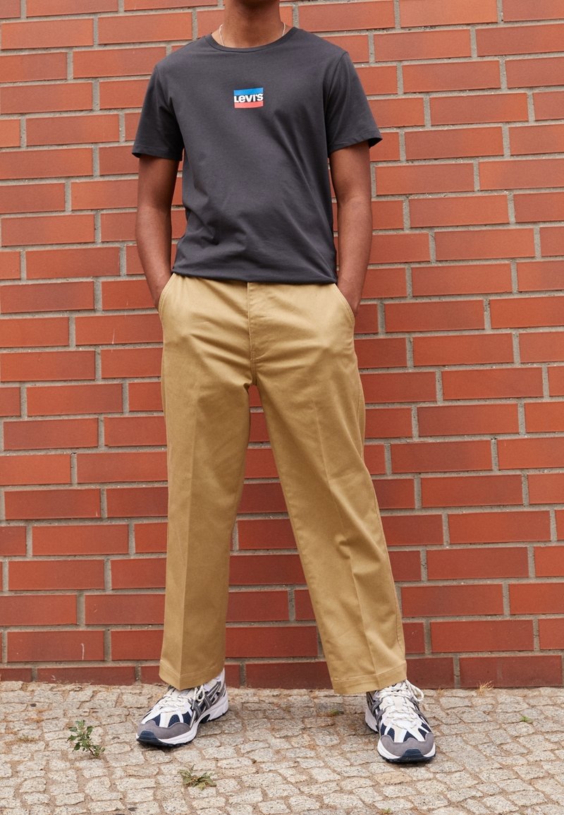 T-shirt en coton gris avec un logo Levi's coloré, associée à un pantalon ample beige et des baskets noir et blanc, se tenant devant un mur en briques.