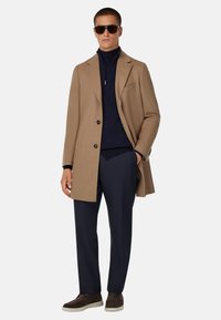 Beige kappa med notch-lapel och två knappar, bärs över en marinblå polo-tröja, ihop med marinblå byxor och mörkbruna slip-on skor.