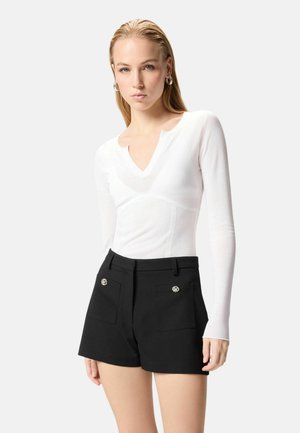 Donna che indossa una maglia bianca a maniche lunghe con scollo a V e pantaloncini neri a vita alta con tasche frontali decorate.