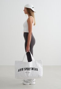 Jonge vrouw in een witte tanktop, zwarte legging en witte pet houdt een grote witte totebag met de tekst "AIM'N SPORTSWEAR" vast, staande in profiel.