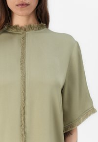 Olivgrünes kurzärmeliges Oberteil aus weichem Material, mit einem Fransenbesatz entlang des Kragens und der Ärmel. Gerader Schnitt.