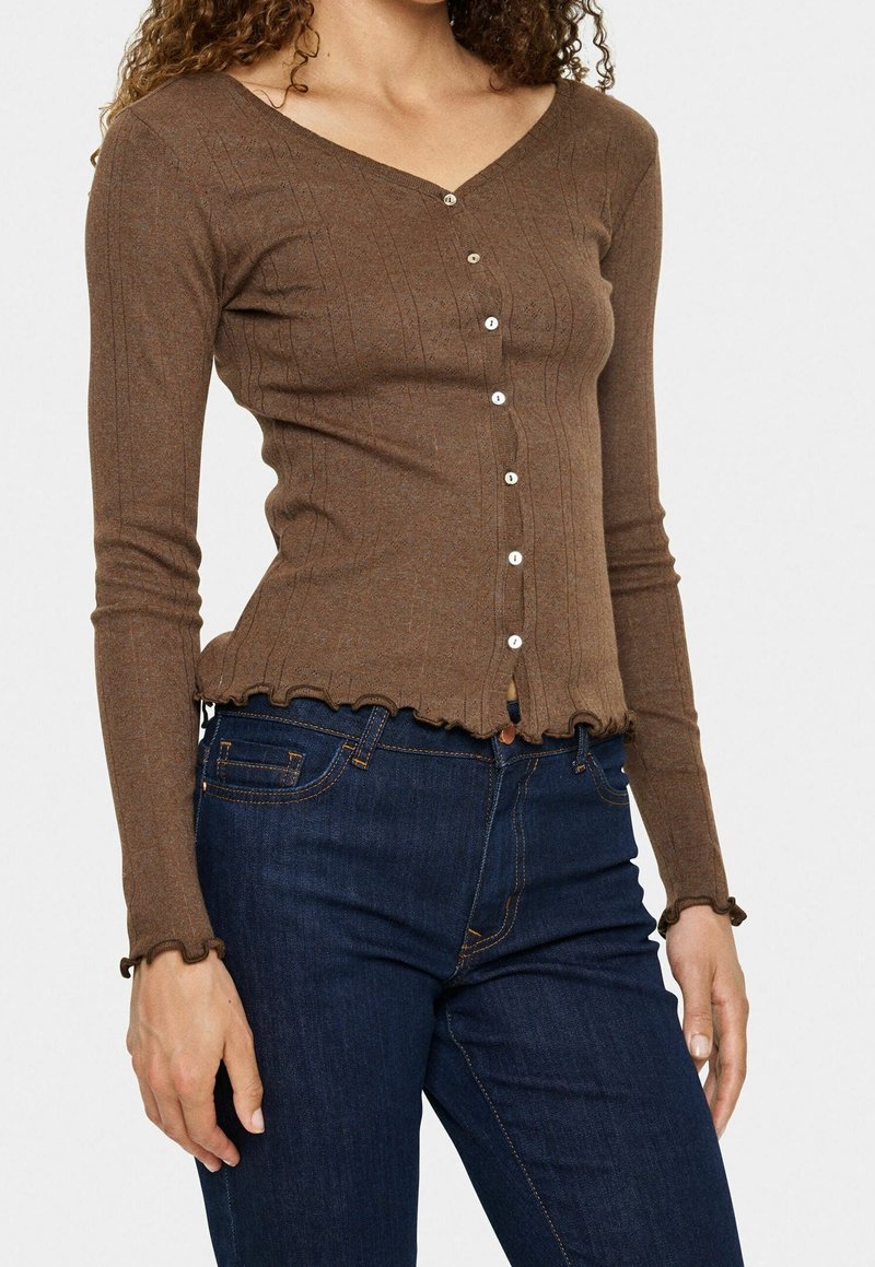 Femme portant un pull ajusté marron à manches longues boutonné avec des bords volantés et un jean bleu foncé, debout devant un fond uni.