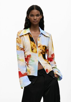 Femme portant un chemisier zippé à fleurs multicolores avec un col oversize et un pantalon noir, debout les mains dans les poches sur fond blanc.