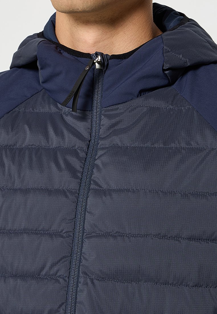 Veste isolée bleu marine avec fermeture éclair, présentant des coutures matelassées horizontales, une capuche et un tissu contrastant sur les épaules.