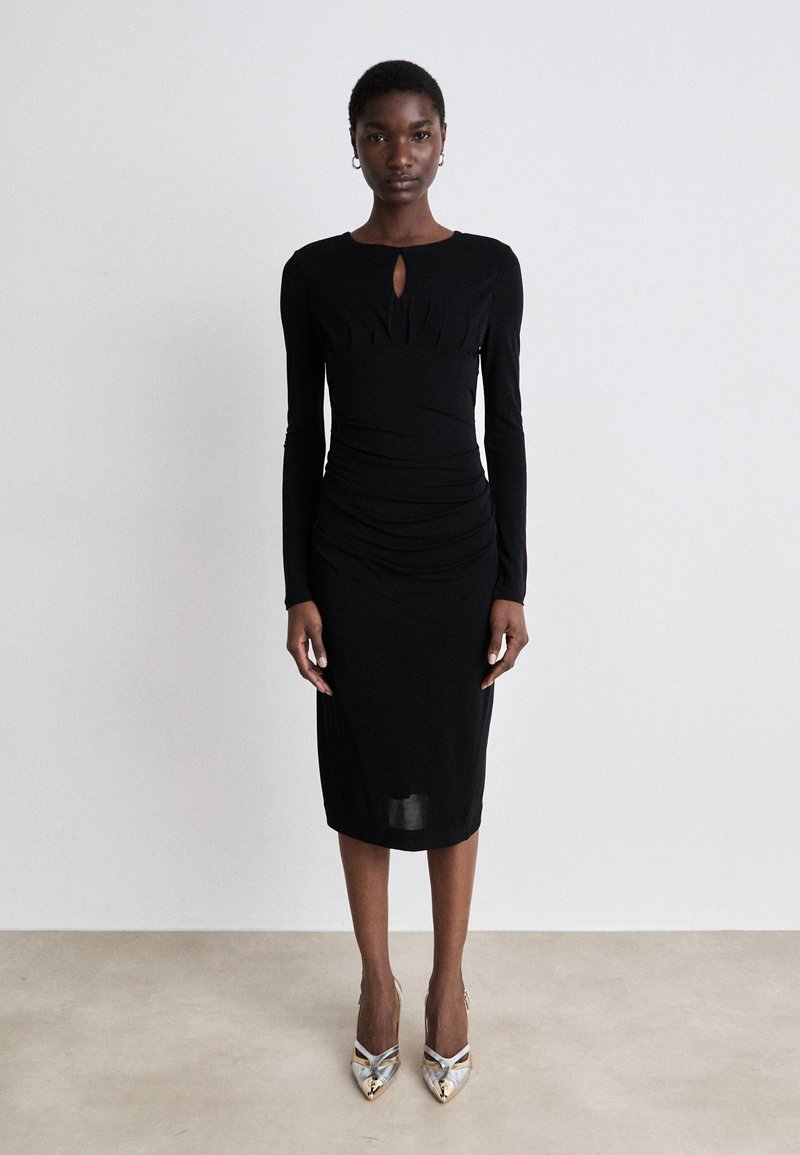 Black long-sleeve dress with a keyhole neckline, ruched texture, and a fitted silhouette, worn with metallic pointed-toe heels.