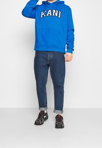 Sudadera con capucha azul con "KANI" impreso en blanco, combinada con jeans de mezclilla ajustados de color azul oscuro y zapatillas negras con detalles en rojo.