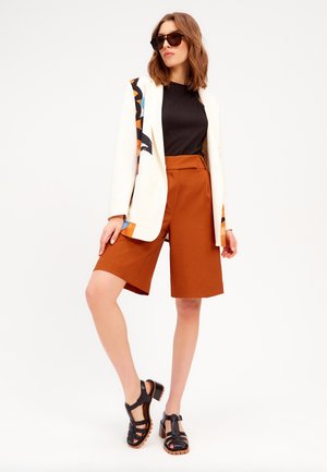 Witte blazer met kleurrijke patroondetails, zwarte top, roestkleurige shorts en zwarte sandaaltjes met hakken. Glad materiaal en gestructureerde pasvorm.