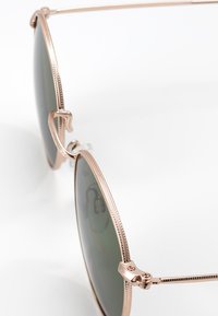 Runde solbriller med ramme i roseguld metal, grøntonede linser og teksturerede detaljer på kanterne af rammen. Slanke wire-arme forbinder til linserne.