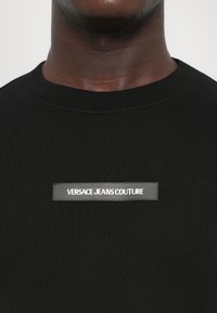 Svart strikkegenser med ribbestrikket rund hals og et rektangulært logo-merke som leser "VERSACE JEANS COUTURE" i hvit skrift.