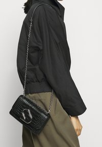 Černá texturovaná kožená crossbody taška se stříbrným kováním a řetězovým popruhem, s klopovým uzávěrem a embosovaným vzorem.
