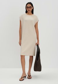 Beige, knielanges Kleid mit kurzen Ärmeln und strukturierter Stoffqualität. Das Model hält eine dunkle, geflochtene Tasche. Schlichte, lockere Passform.