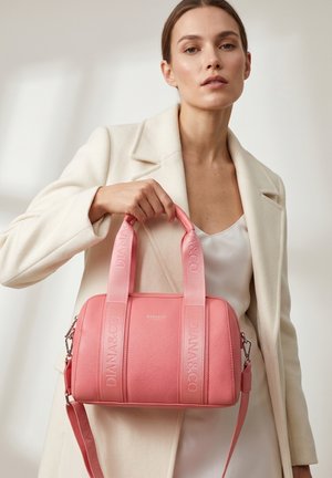 Femme en manteau blanc tenant un sac à main en cuir rose Diana & Co avec des anses embossées sur un fond neutre.